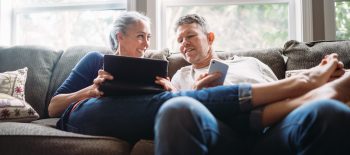 A couple in their 50's relax in their home on the living room couch, enjoying reading and surfing the internet on their mobile touchscreen phones and computer tablet.